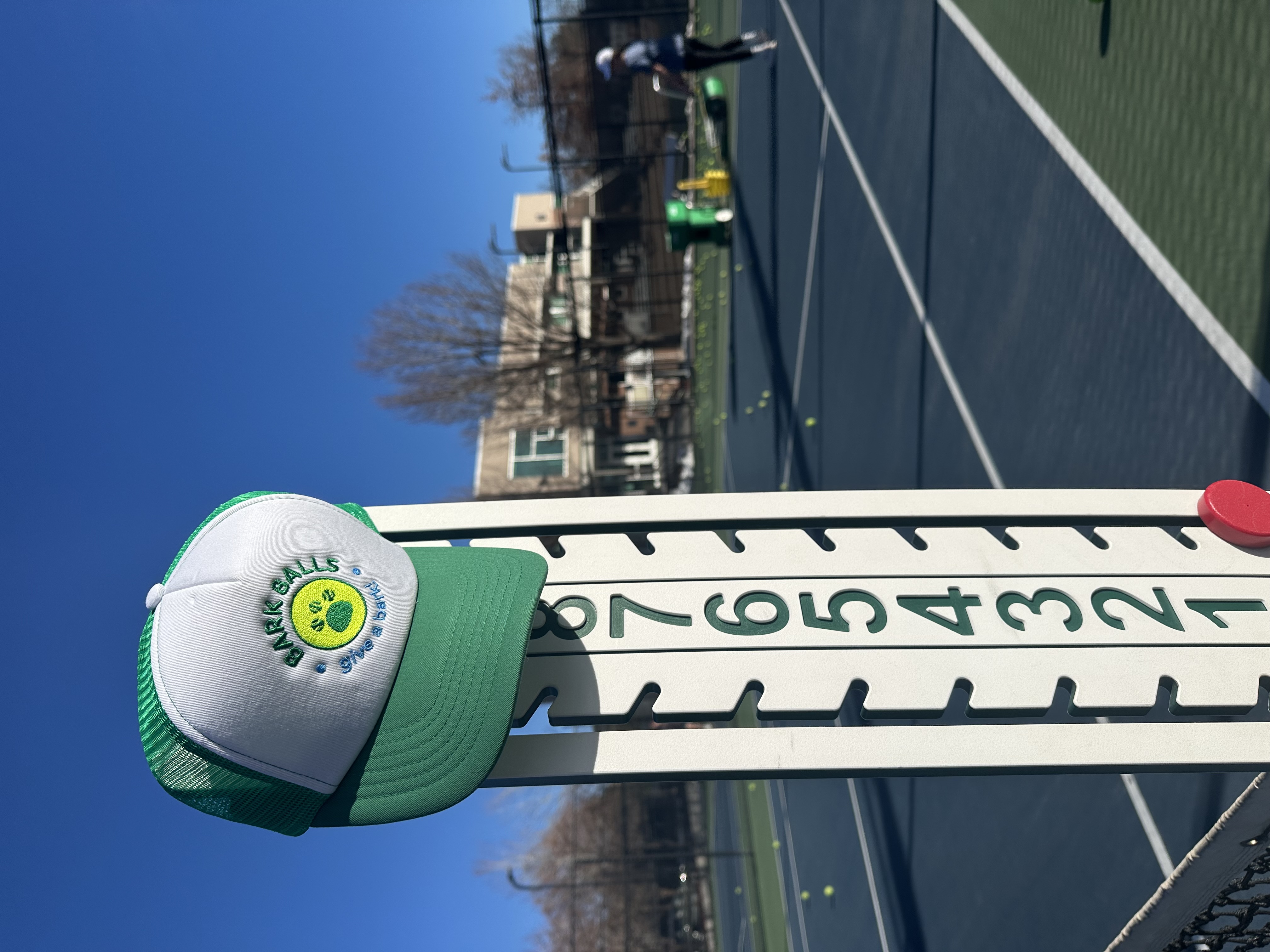 Bark Balls hat on a tennis court scoreboard with player and blue sky in background